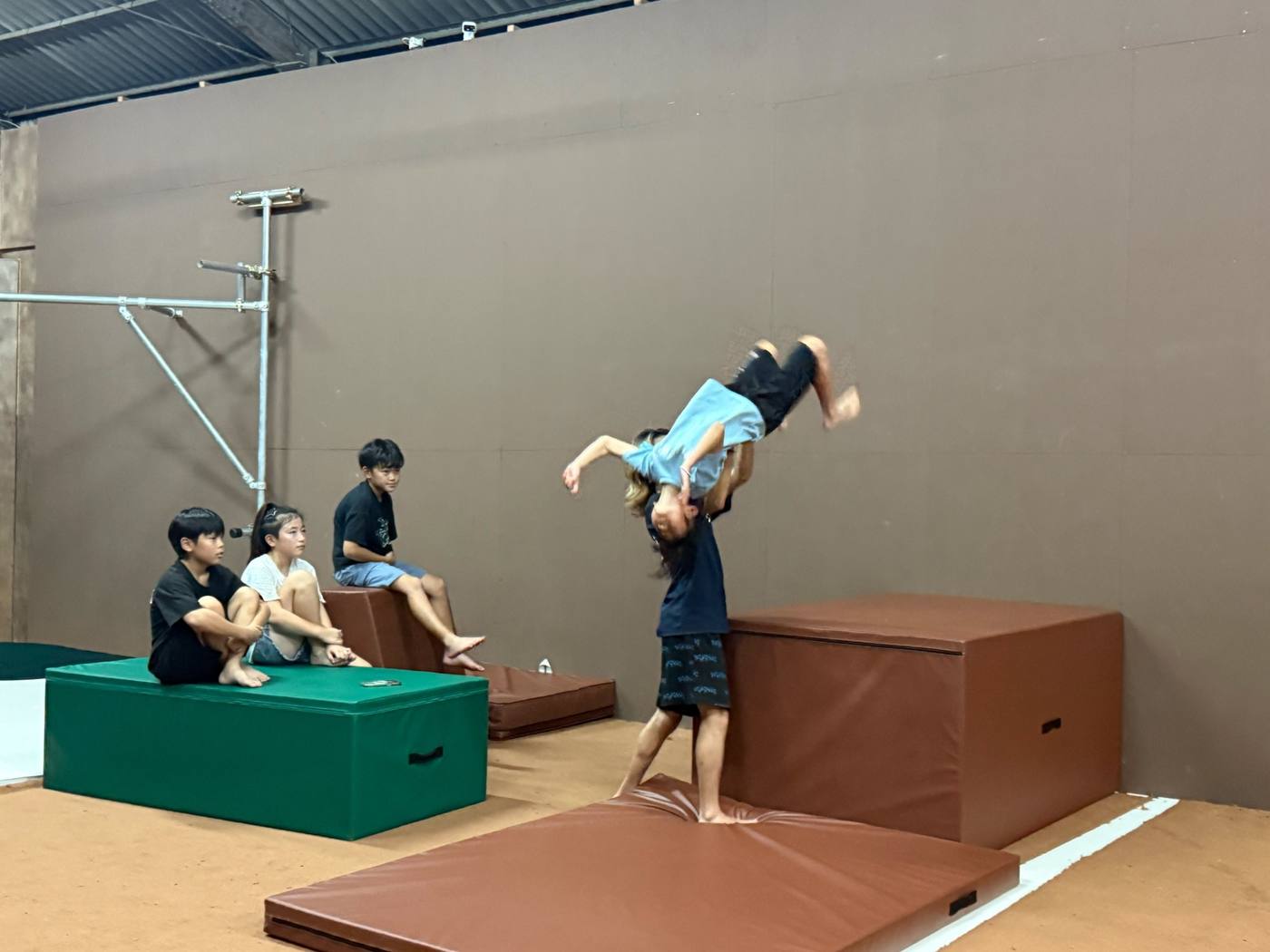 コーチの補助を受けて回転技を練習する子どもと、マットの上で見守る子どもたち